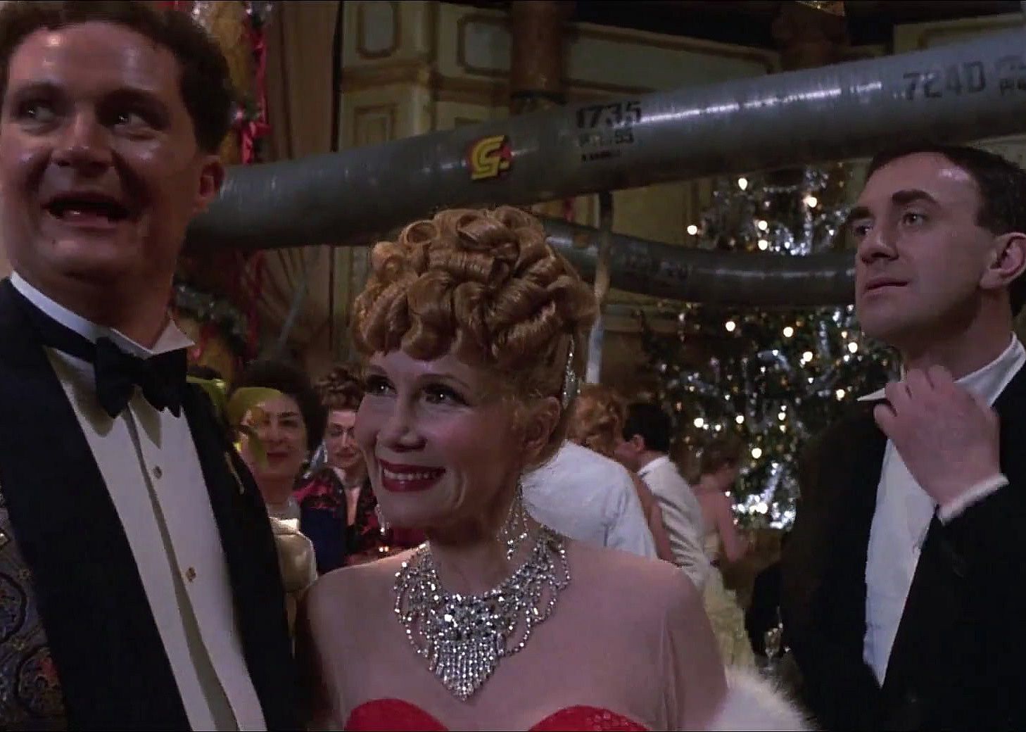 Two men in suits and a redheaded woman in a red gown and jewels at a party.