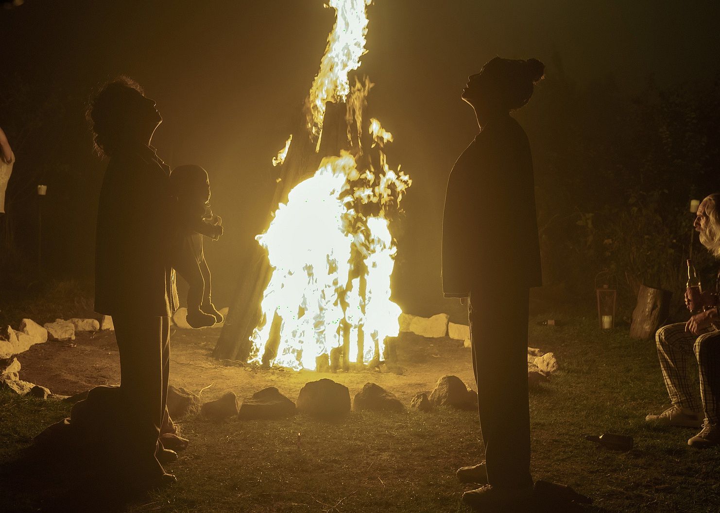 Two women standing next to a bonfire, one of them holding a baby as they look up.