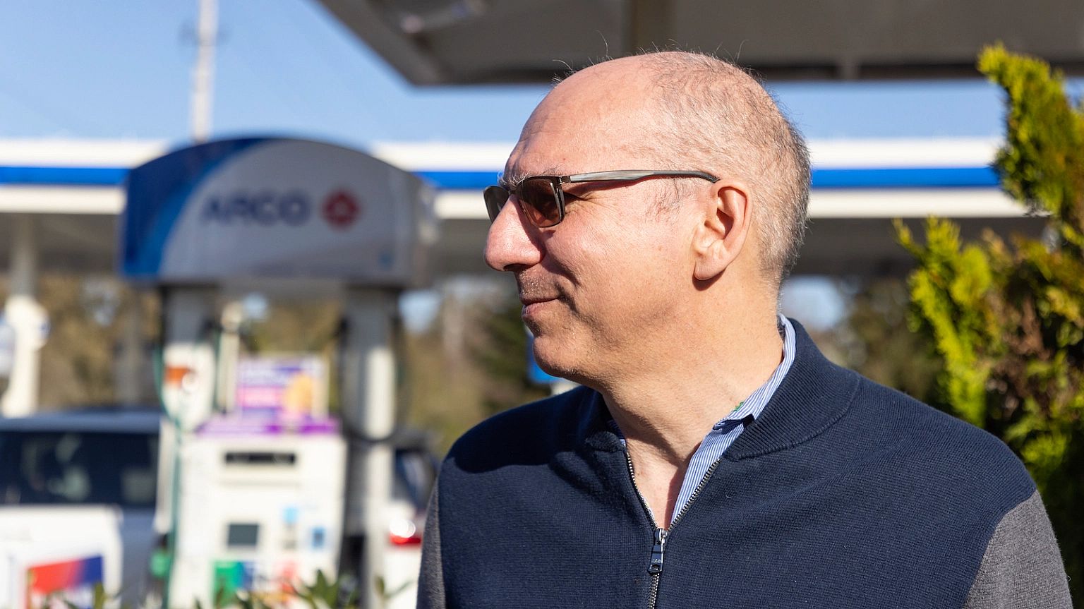 A man wearing sunglasses stands in front of a gas pump.