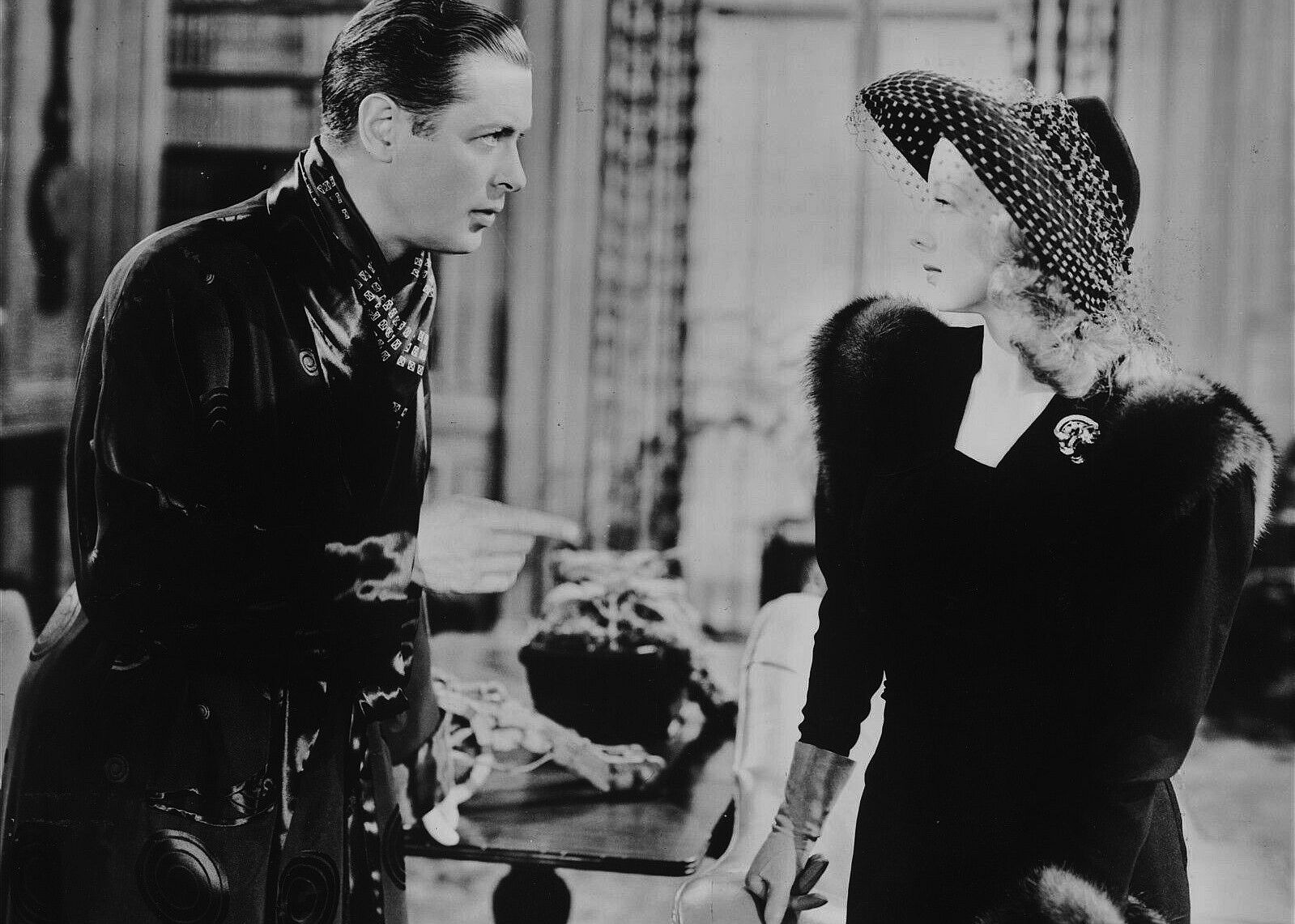 A black and white shot of a handsome man in a silk robe talking with a beautiful woman in a fur coat.
