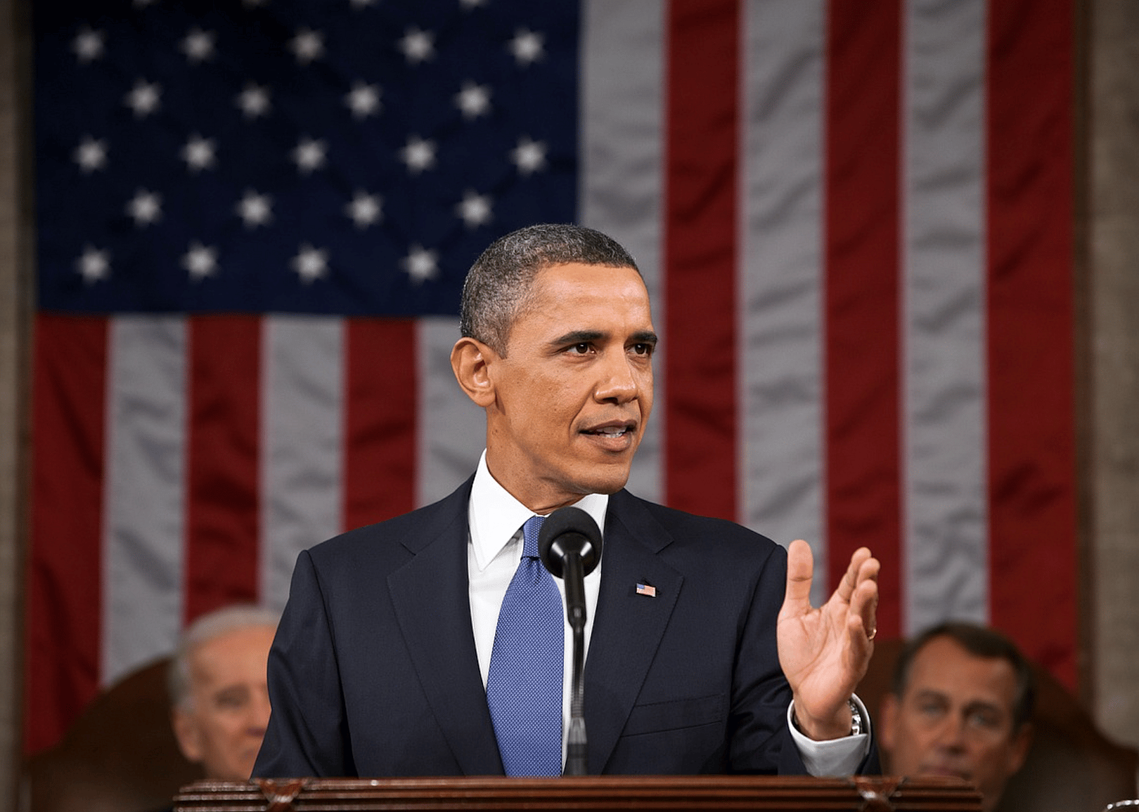 Barack Obama in a black suit at a podium in front of the American flag.