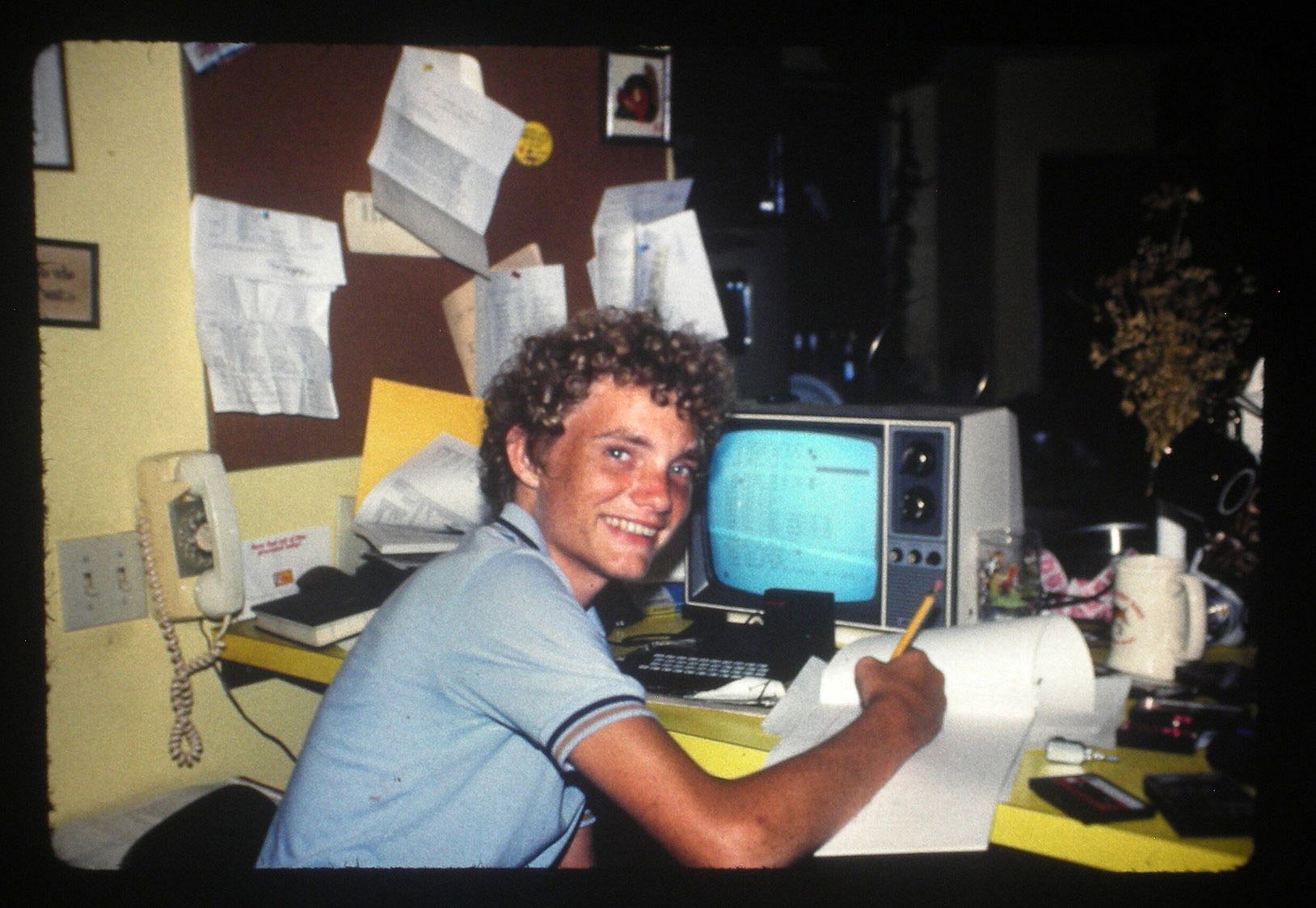 A teenage boy sits at a cluttered desk writing on paper next to a small CRT monitor connected to a Timex Sinclair 1000 computer, 1982.