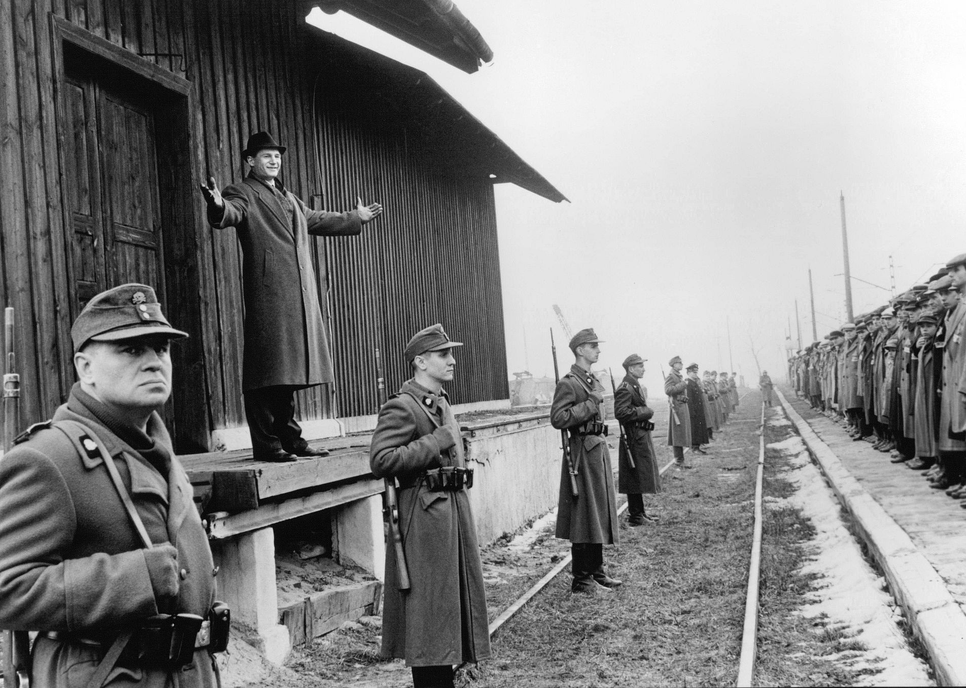Liam Neeson standing on a train platform behind soldiers talking to a line of people.