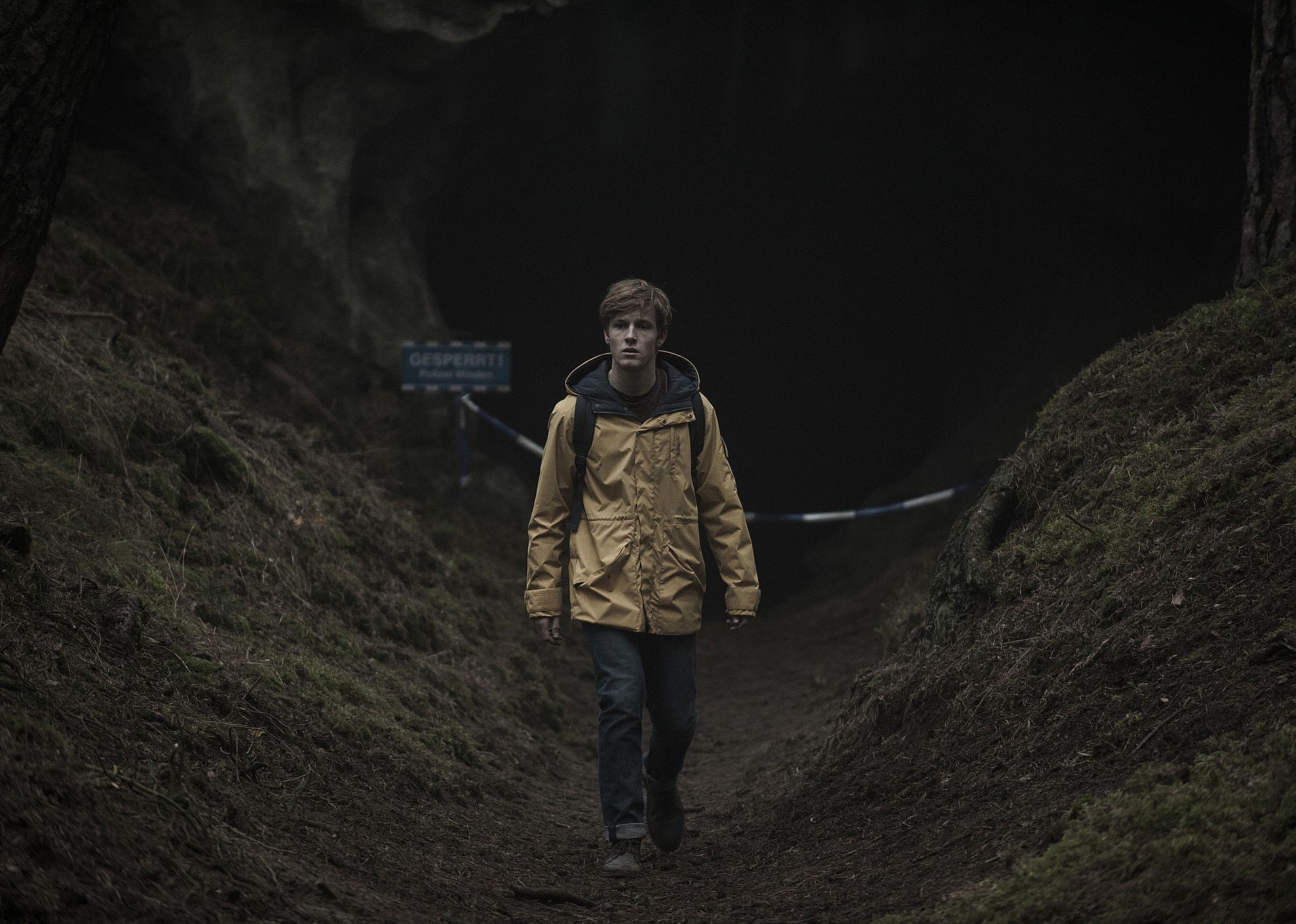 A boy in a yellow raincoat walking near a dark cave.