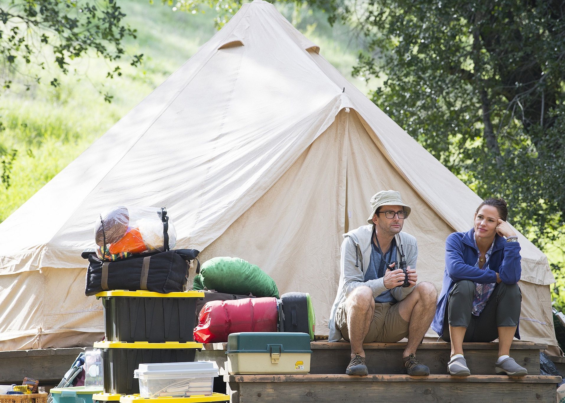 A man and a woman sit on a platform in front of a tent and camping gear.