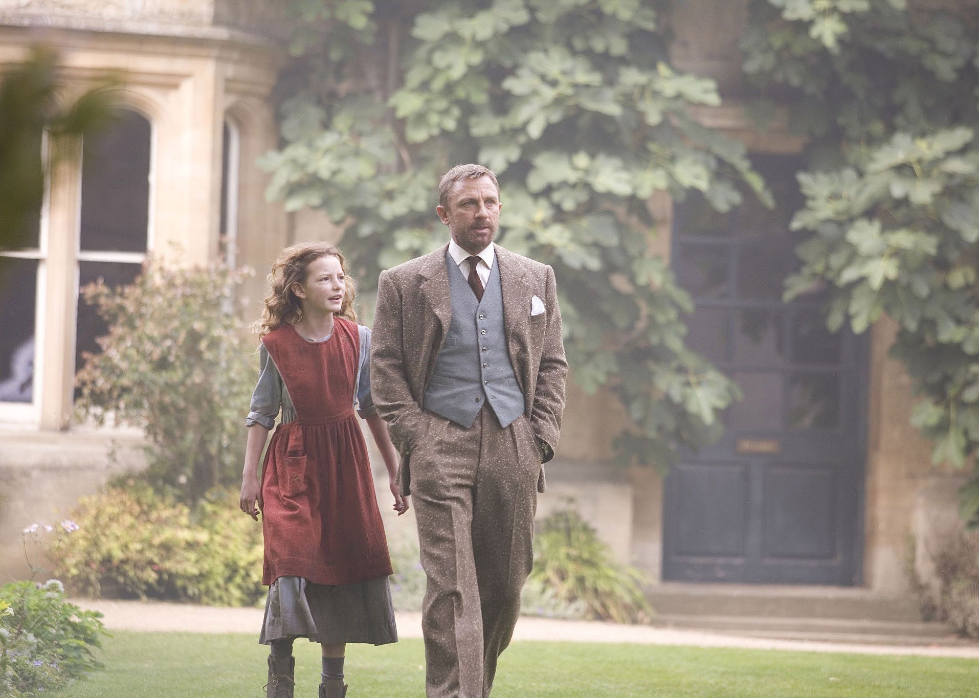 Daniel Craig wears a brown suit and walks with a little blonde girl in a dress and red apron.
