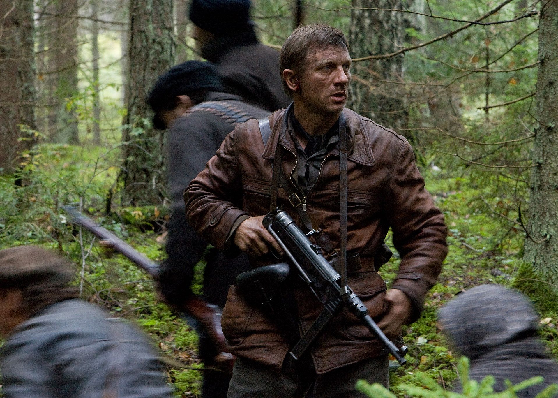 Daniel Craig in a brown leather jacket standing guard with a large gun around his chest as people pass through the woods.