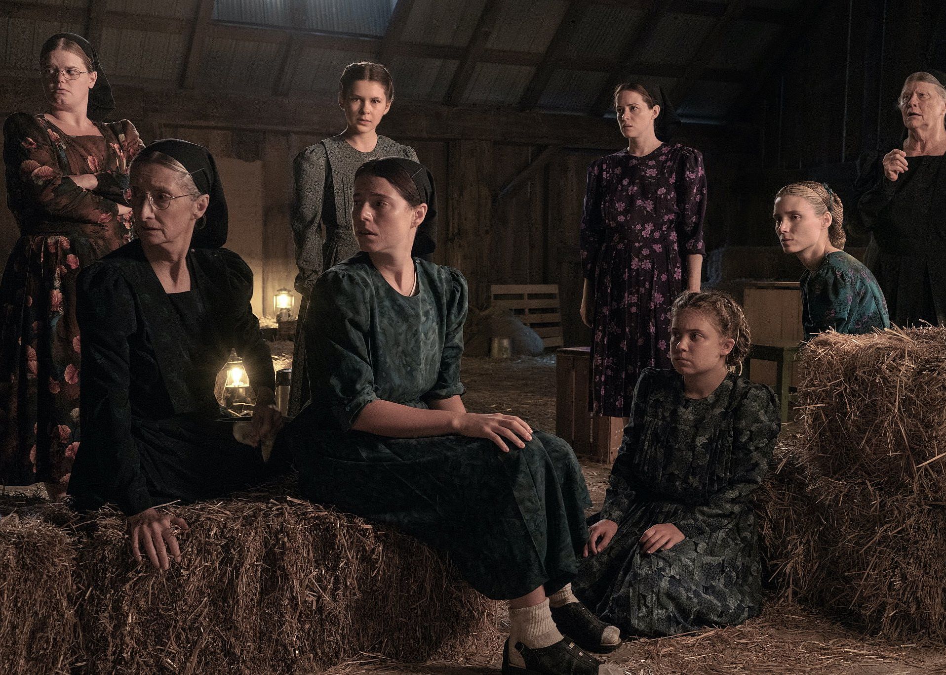 A group of women with worried looks on their faces sit on hay bales in a barn.