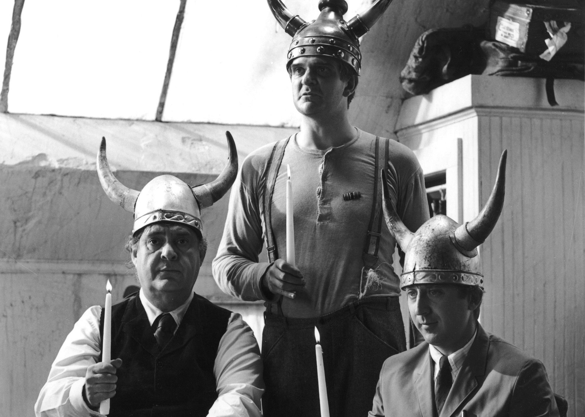 Three men at a table wearing viking helmets and holding candles.