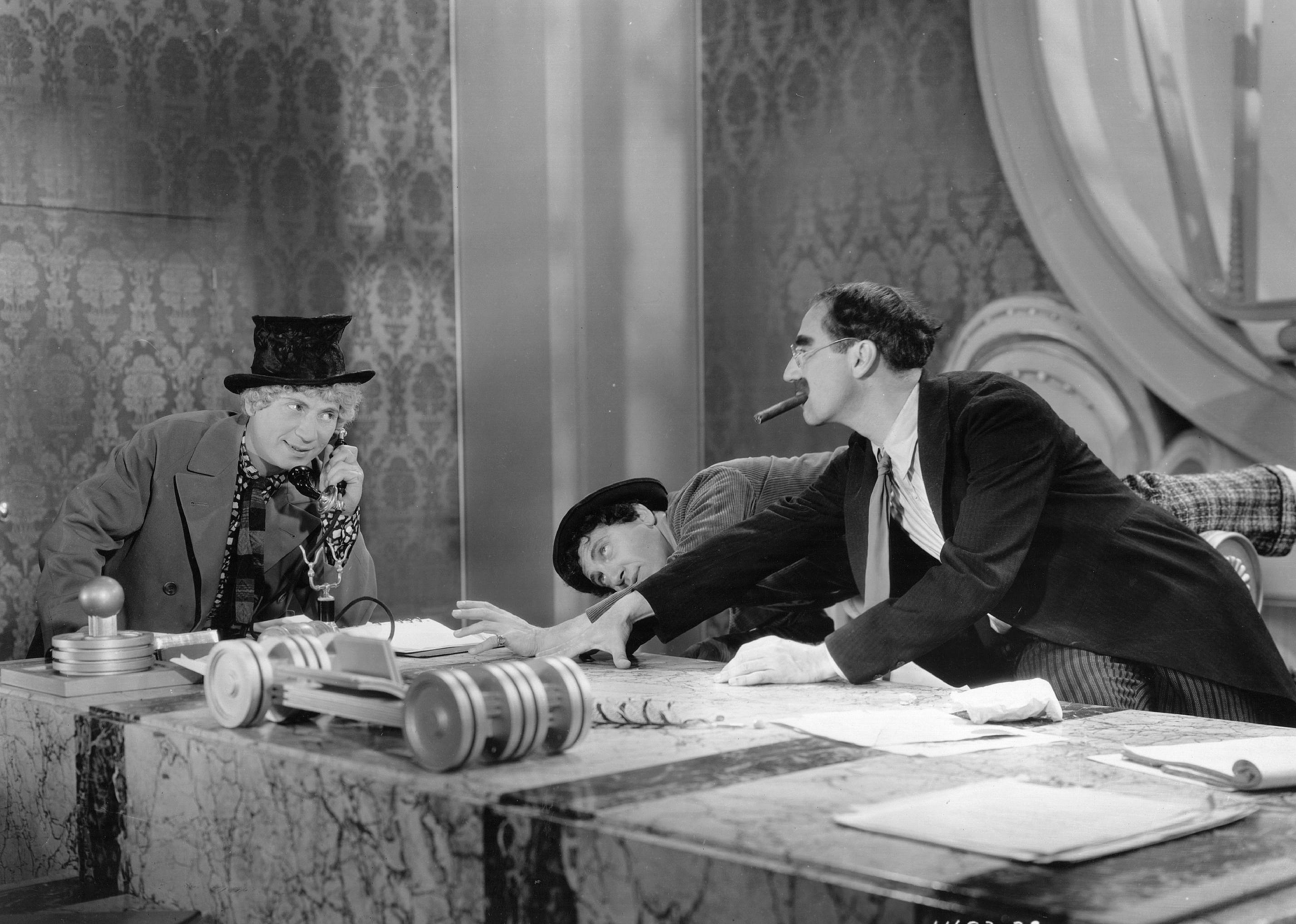 The three Marx brothers at a desk, one on the phone, one looking under and the other on top of the desk smoking a cigar.