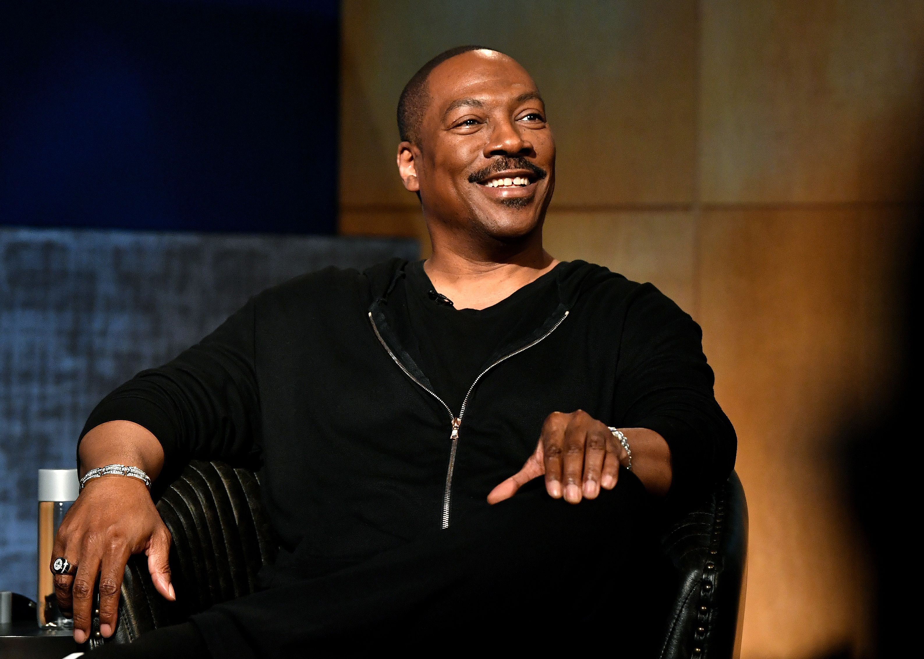 Eddie Murphy in a black hoodie sitting onstage.
