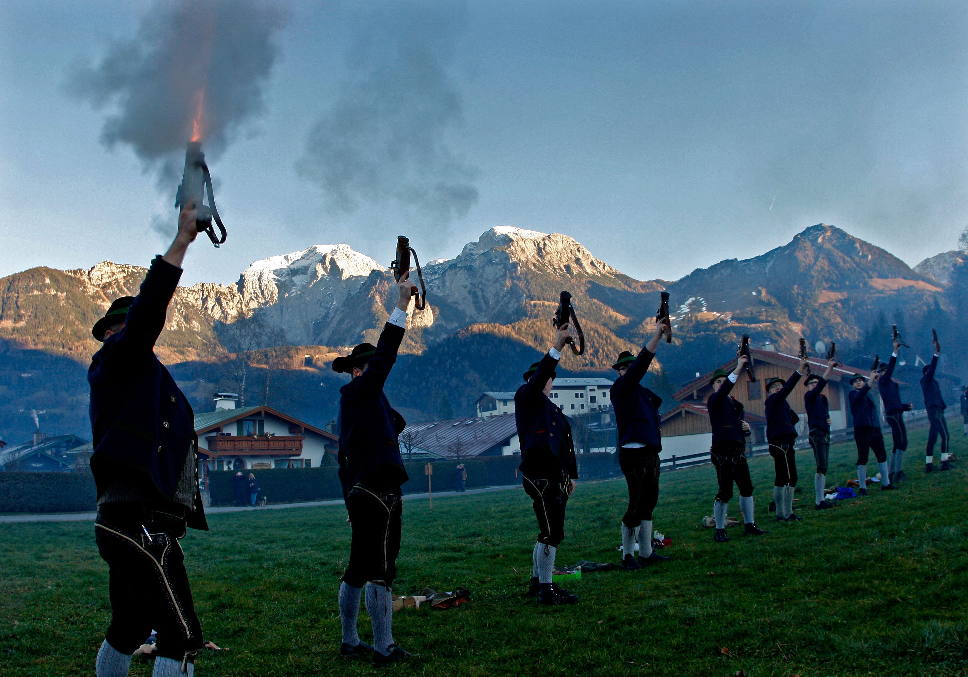 Several men in lederhosen firing mortars into the air with a backdrop of houses and snow capped mountains.