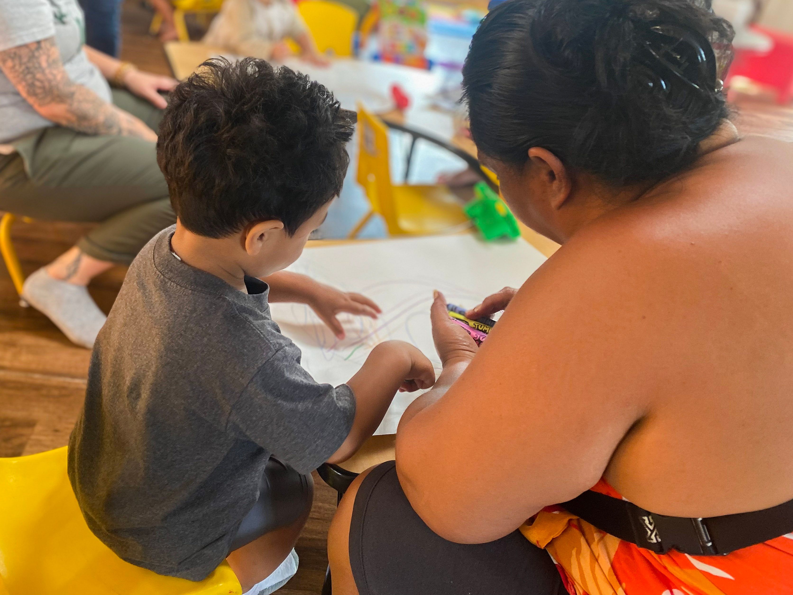 A woman sits with a child while they draw on paper.