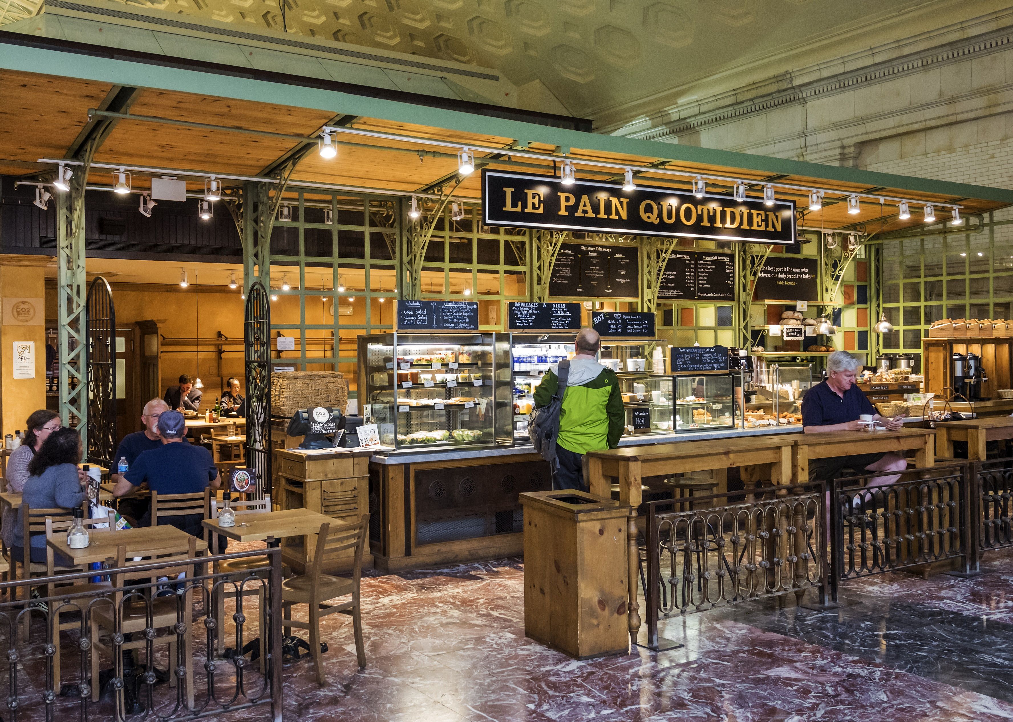 The interior of a Le Pain Quotidien bakery with a customer waiting to be served while other customers sit in. 