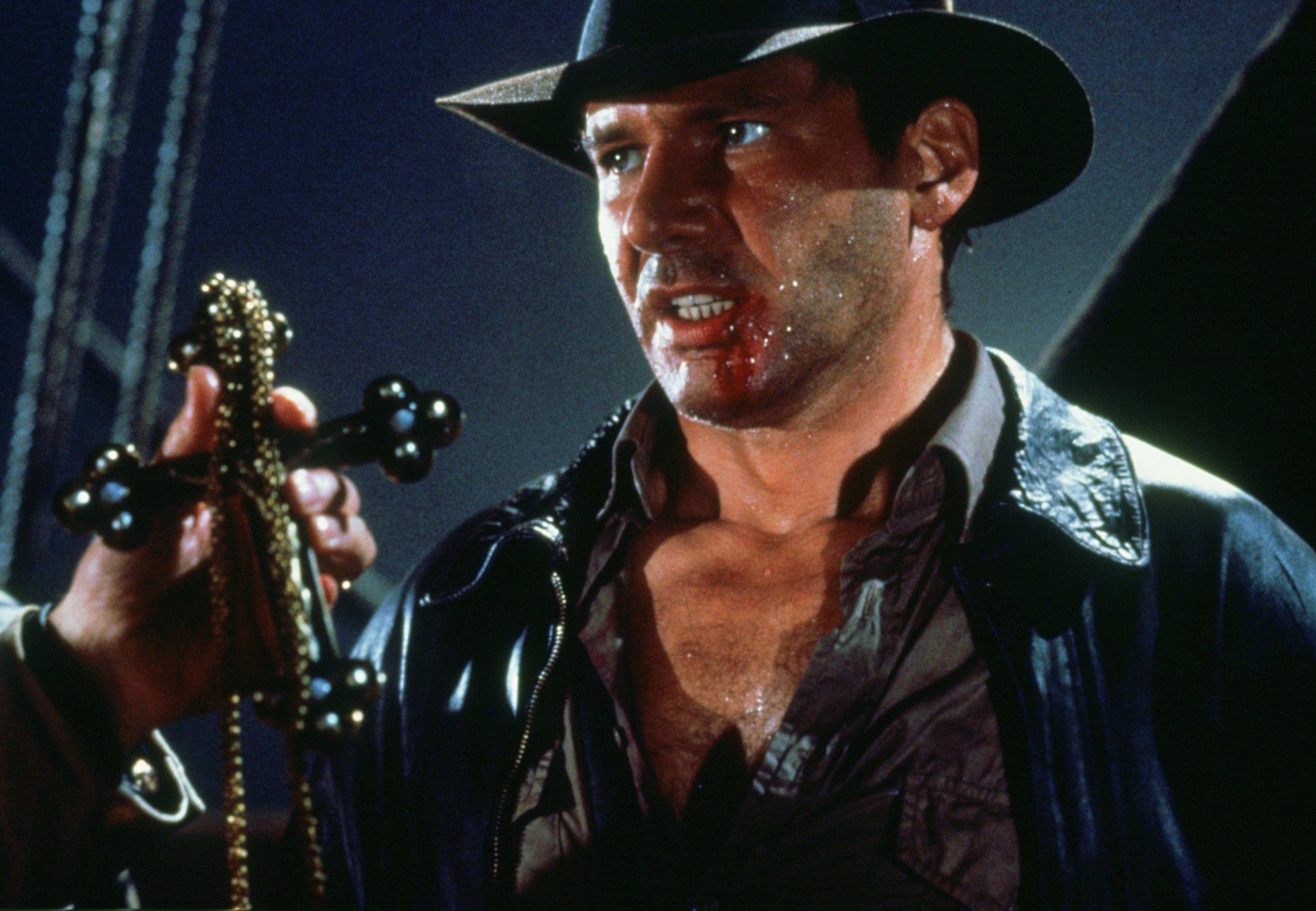 Harrison Ford in a hat and black leather jacket standing in front of someone holding a cross.