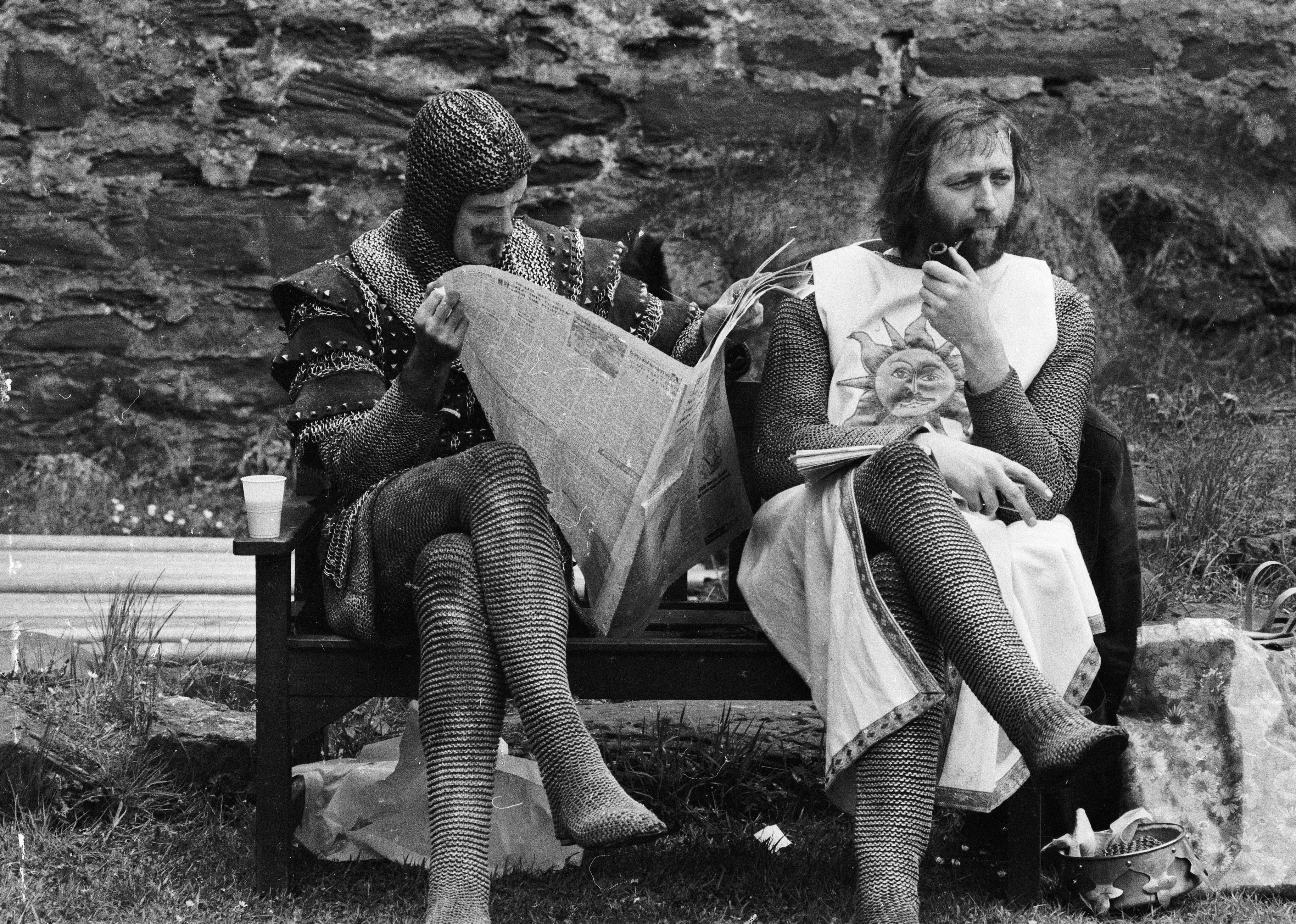 Two knights sitting on a park bench, one reading the paper and the other smoking a pipe.