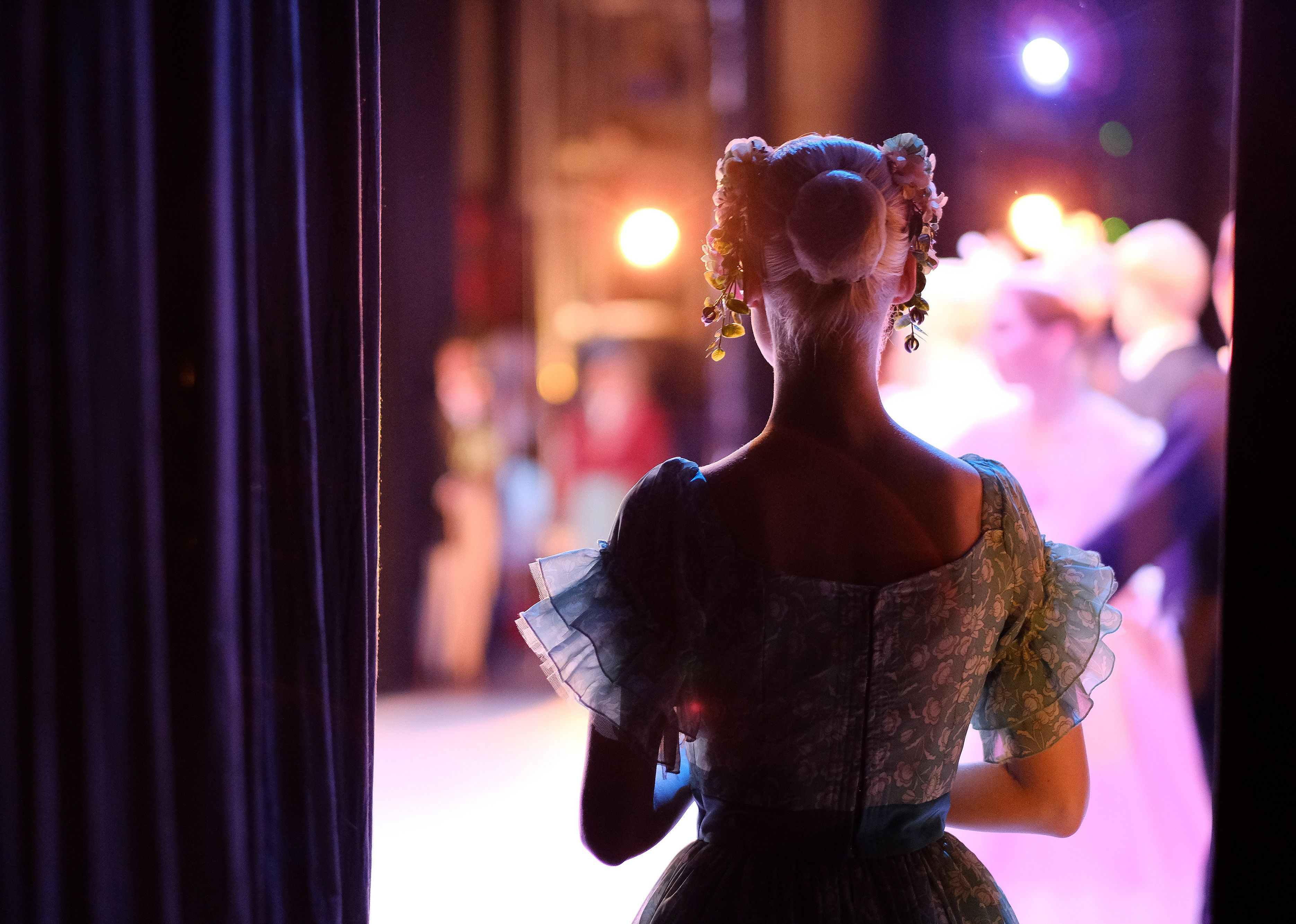 Stage actress in costume watches performers through curtain.