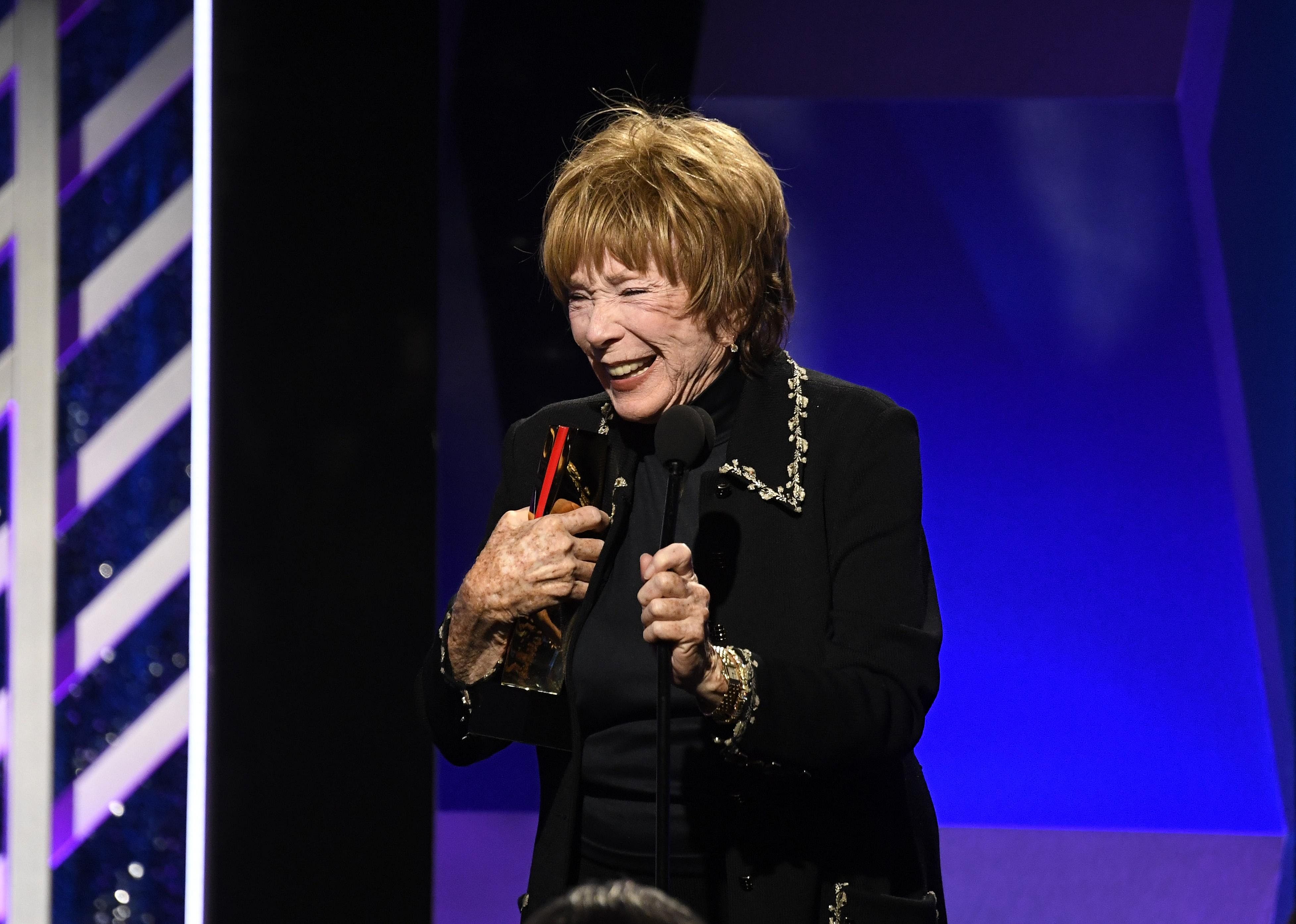 Shirley MacLaine smiling and speaking into a mic on stage.