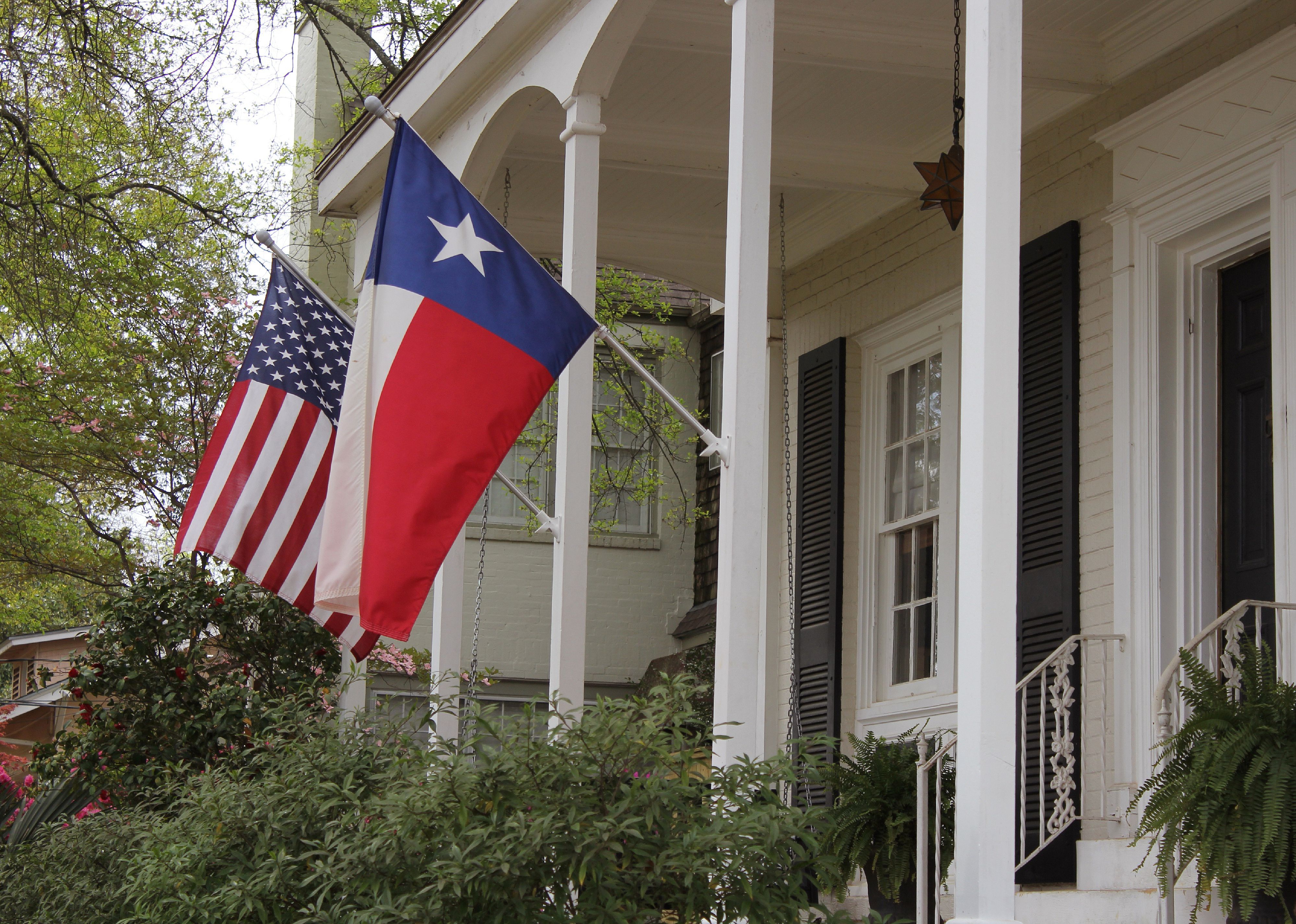 A large white home with dark shutters and a U.S. flag and a Texas flag flying in front.