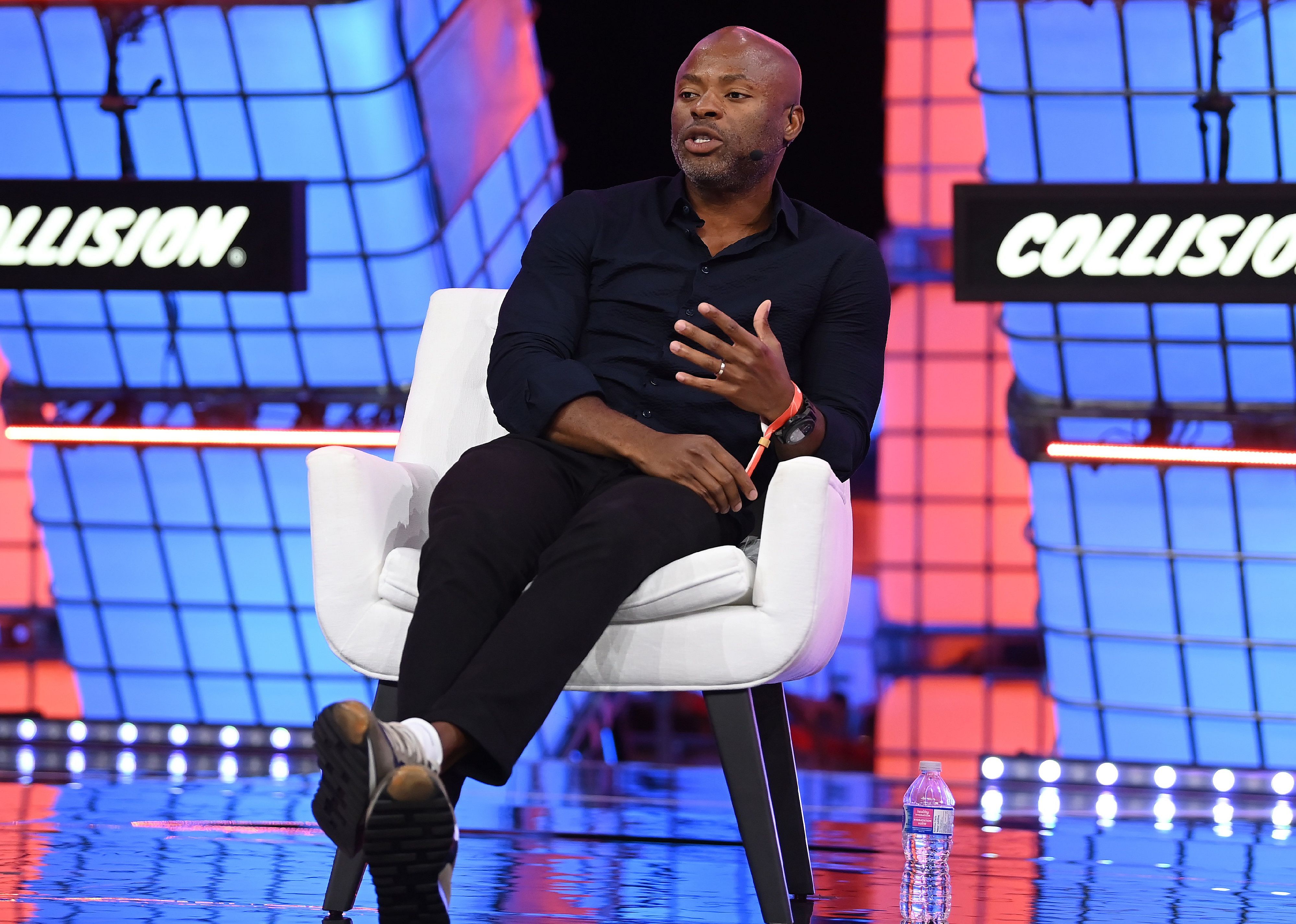 Tope Awotona speaking onstage in front of a red and blue background.