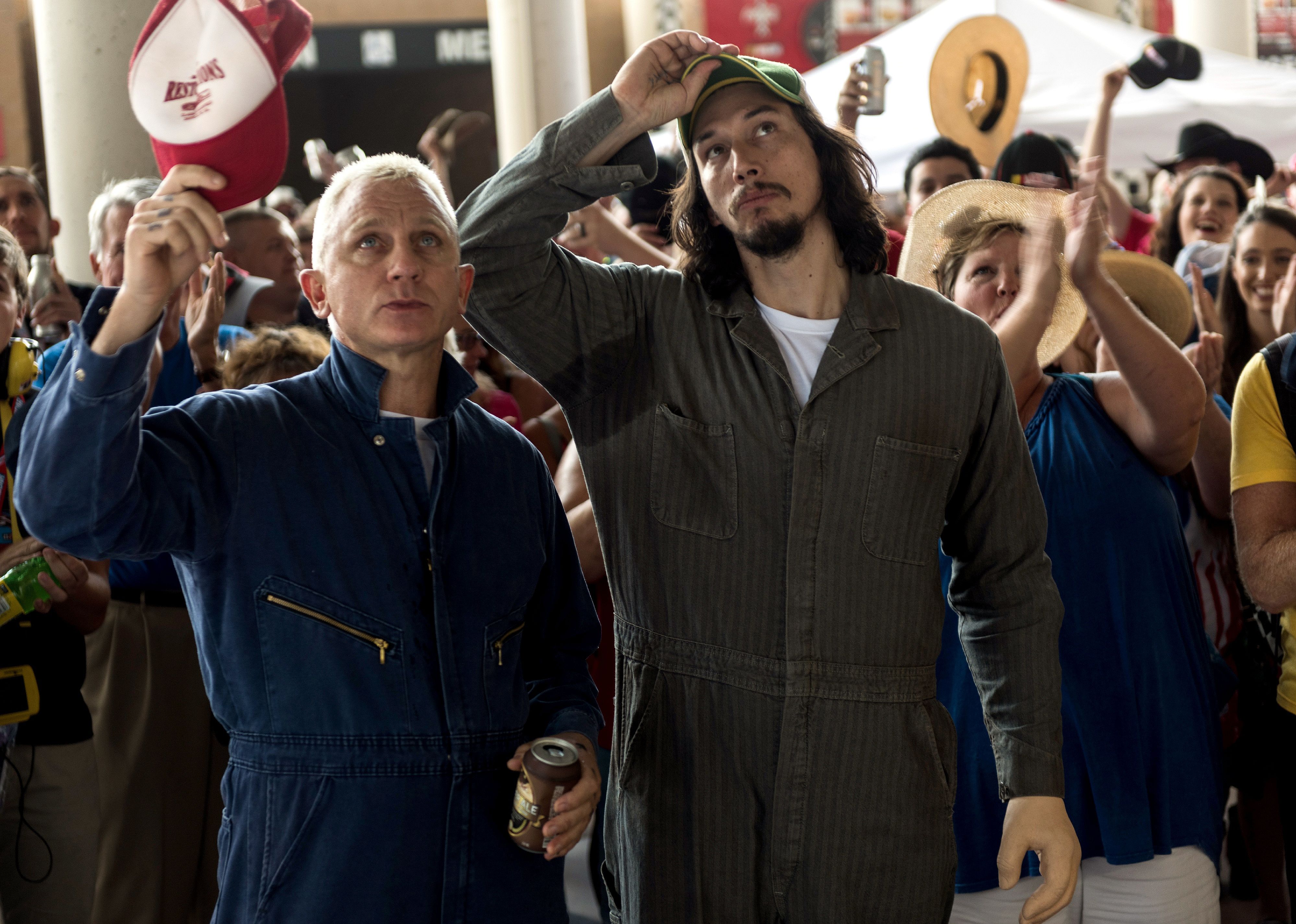 Daniel Craig and Adam Driver in mechanic uniforms at a car race.
