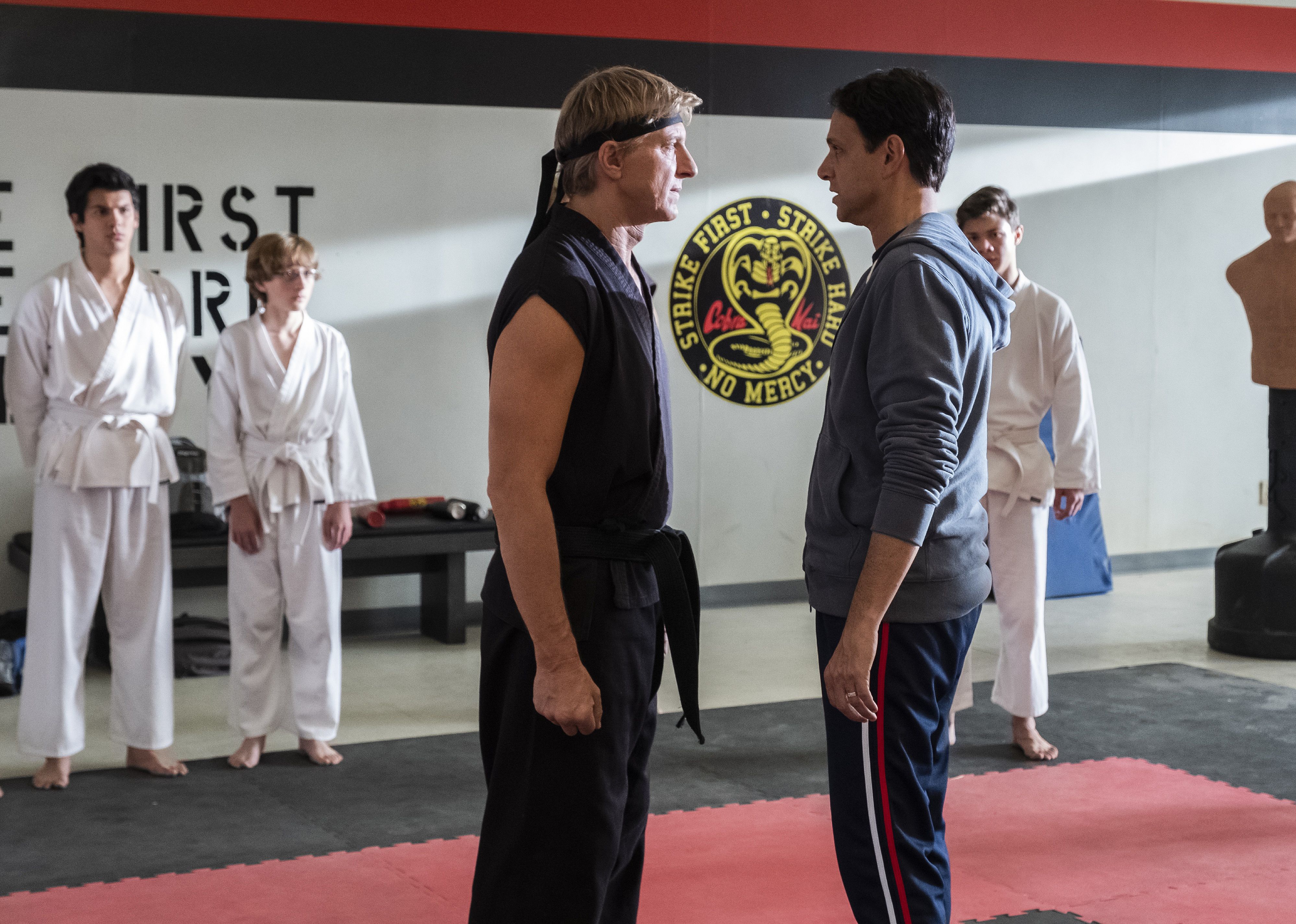 A man in black karate gear stands facing a man in a hoodie and sweat pants in front of kids in white karate clothes.