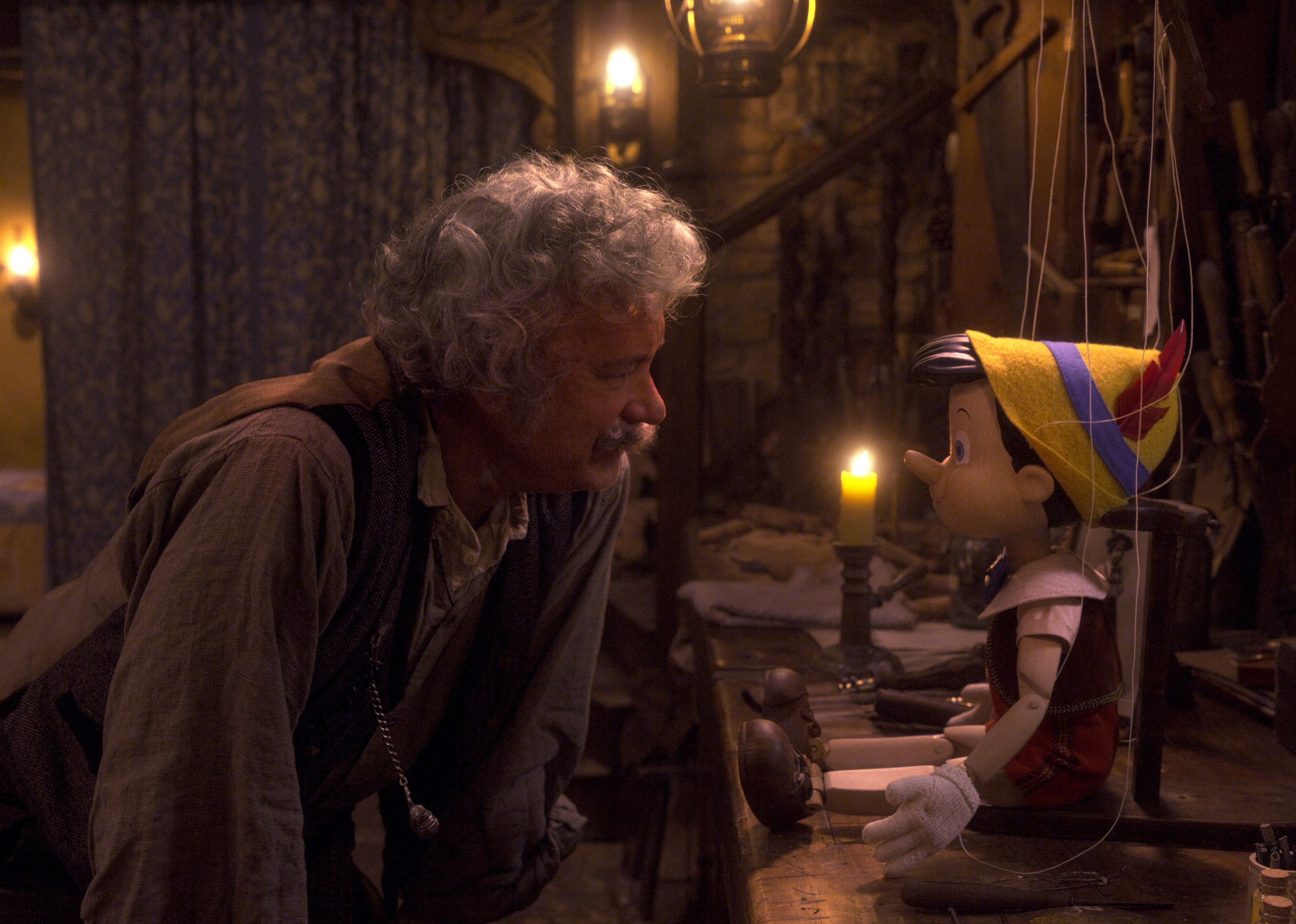 An older man (Tom Hanks as Geppetto) leaning over a table to look at a puppet of a little boy (Pinocchio).