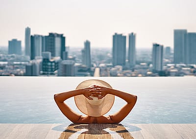 Woman wearing hat in urban infinity pool, looking out toward city buildings.