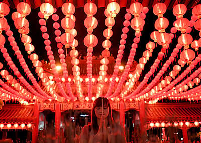 Red lanterns line the ceiling above a woman with her eyes closed and her hands in prayer.