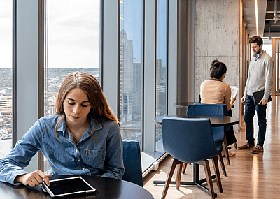 A young woman is seated by an office window with other employees in the background.