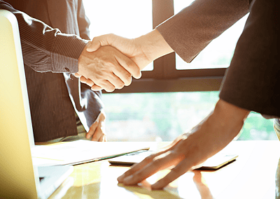 A handshake takes place over a desk and a laptop.