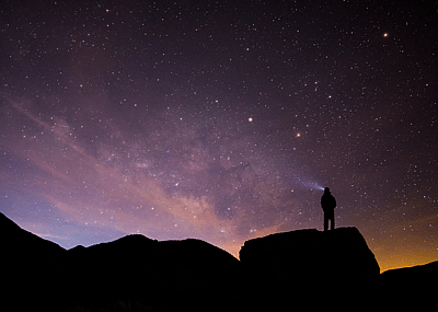 Silhouetted person stands atop boulder with light beam looking up at the milky way and sunrise.