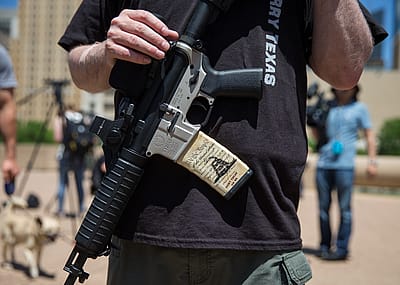Person with an assault rifle across their chest, a miniature copy of the American constitution on the gun handle, at a counter-protest by gun advocates in response to protesters opposing the NRA's annual convention in Dallas, Texas, 2018.