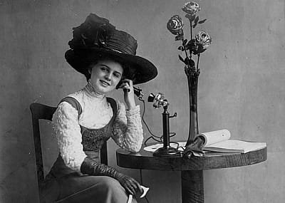 Person with hat seated at table talking on an early standing telephone.