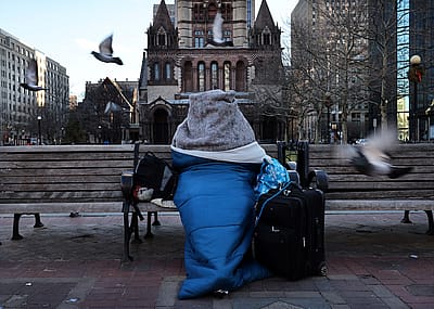 Bundled in blankets and a sleeping bag, a homeless person sits on a bench in Copley Square in Boston.