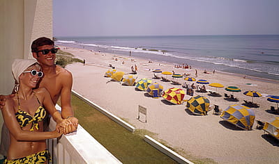 A smiling couple lean on a balcony at Thunderbird Motel, which overlooks a section of beach shaded by various colored umbrellas, Myrtle Beach, South Carolina, 1960s.