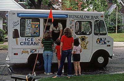 View of a small group of children and teenagers at a Bungalow Bar-brand ice cream truck on Fitchett Street (near 64th Road), in the Rego Park neighborhood, Queens, New York, New York, July 1981.