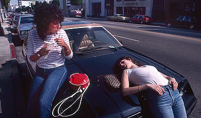 Two teenage girls sit and lie on the hood of a parked car on a Beverly Hills street, one eating from a red thermos cup, 1982.