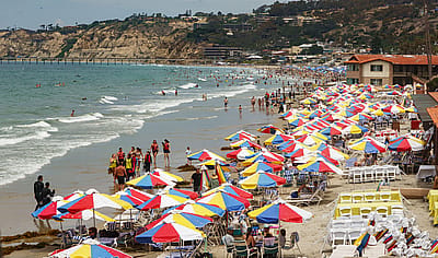 Rows of colorful umbrellas fill a packed beach with swimmers and surfers in the water, capturing a classic summer day at the coast.