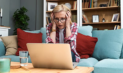 An older woman holding her head in shock as she looks at a laptop.