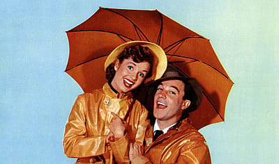 Lobby card artwork for “Singin’ in the Rain” (1952) showing Debbie Reynolds and Gene Kelly smiling under a large orange umbrella against a pale blue background. Both wear shiny yellow raincoats; she has a yellow rain hat and he wears a brown fedora.