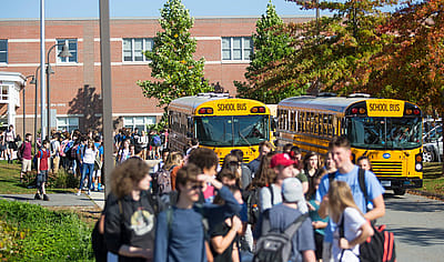 High school students walk along a sidewalk outside a brick school building as two yellow school buses wait under trees with fall foliage.