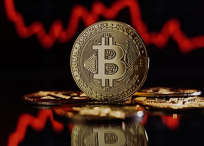 A physical representation of a bitcoin, with the B symbol prominently displayed in the center of the coin, in front of a background of a red, downward-trending line graph.