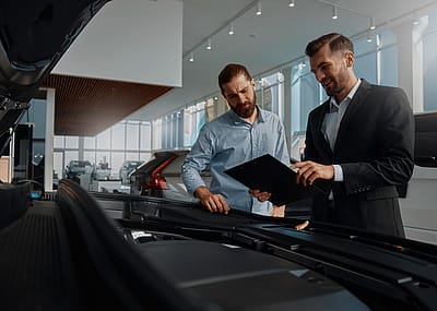 Two men standing in a car dealership or showroom. One man dressed in a suit, is holding a clipboard showing something to the other person, who is casually dressed. They are both looking at the open hood of a car.