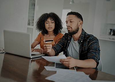 A young couple sitting at the table with a laptop, papers, and a credit card in front of them.