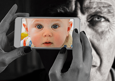 A close up of hands holding a cell phone with an image of a baby on the screen with an older man image in the background.