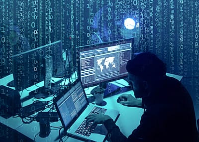 A man's silhouette in a dark room sitting in front of a laptop and another computer screen.