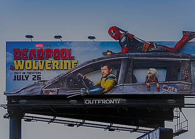 A billboard advertisement promoting the upcoming movie Deadpool & Wolverine by Marvel Studios, set to release on July 26. There is a man dressed in black and red bodysuit lying on top of a damaged car, while a man is in the driver's seat looking focused and intense.