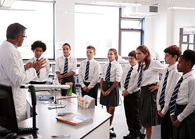 A male educator instructing a group of high school students dressed in their school uniforms, huddled around his desk.