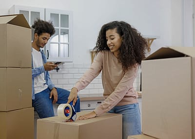 A young couple in a kitchen surrounded by boxes. A woman is holding a packing tape and taping a large box on the floor while a man is sitting down on the counter next to a large pile of boxes looking at a phone in his hand.